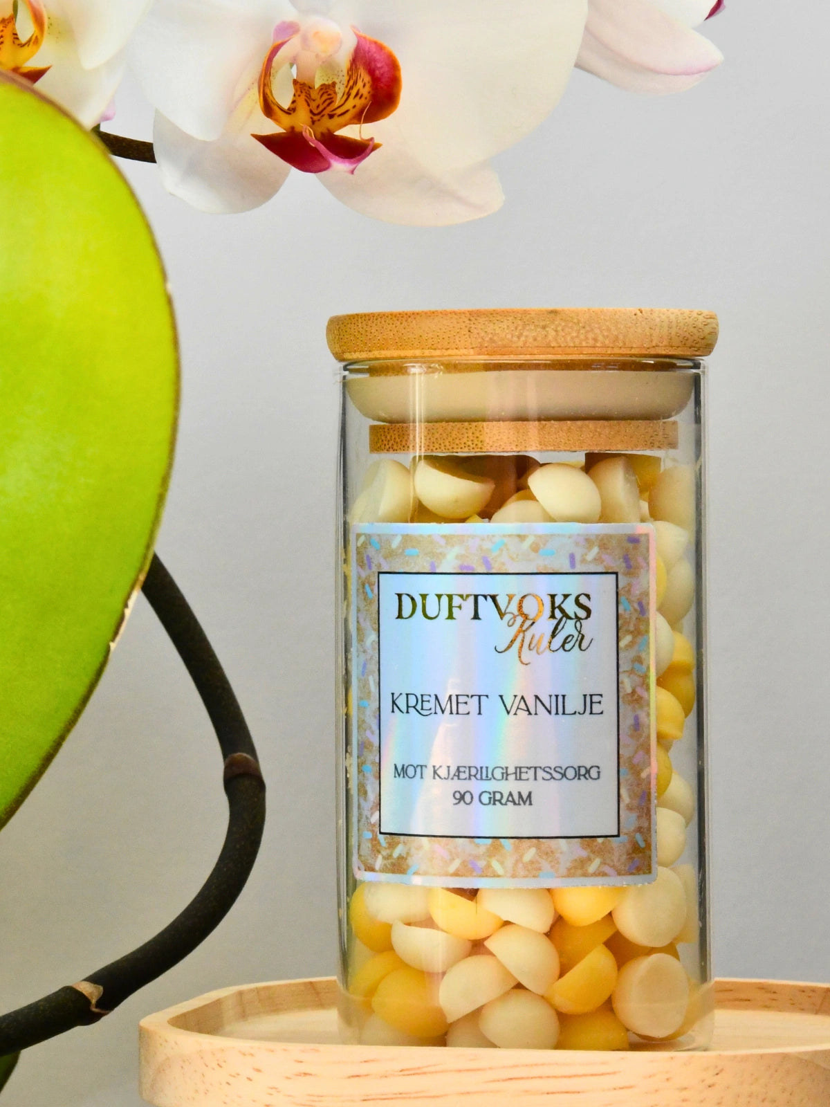 Kremet vanilje duftvoks som er håndlaget i Norge. På bilde står den i sin glasskrukke med bambus lokk og en nydelig ettikkett. Over er en orkide. Denne duftvoksen er perfekt gave til noen som har kjærlighetssorg. Fyll rommet med en herlig duft av kremet vanilje og se lyst på fremtiden. Drømmepartneren din er der ute en plass, bare vent å se!