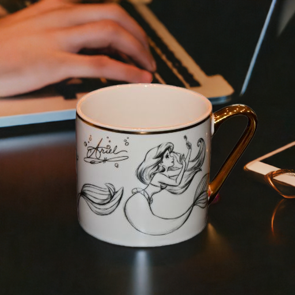 Ariel fra Disney. Kopp med bilde og tekst. Gull detaljer. Her står koppen på et bord mens noen sitter å jobber på en PC.