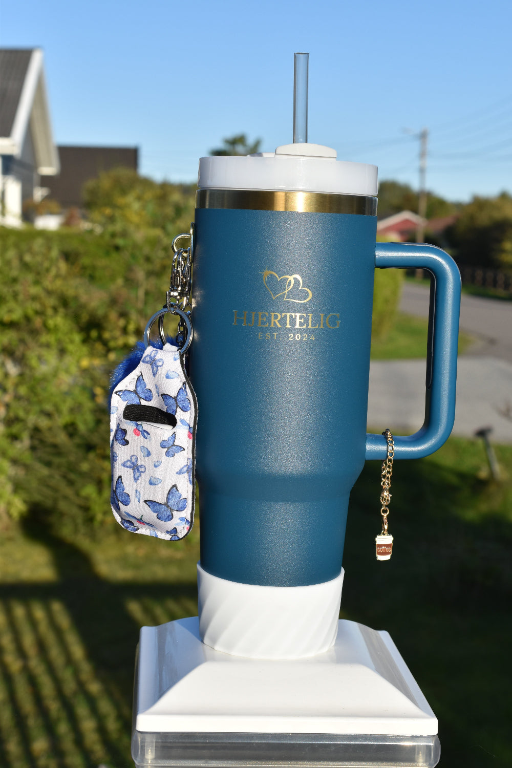 Blå termokopp med Hjertelig logo og gullkant, pyntet med tilbehør som silikonbunn, smykke med kaffemotiv rundt håndtaket, en blå dusk og en veske til airpods med blå sommerfugler
