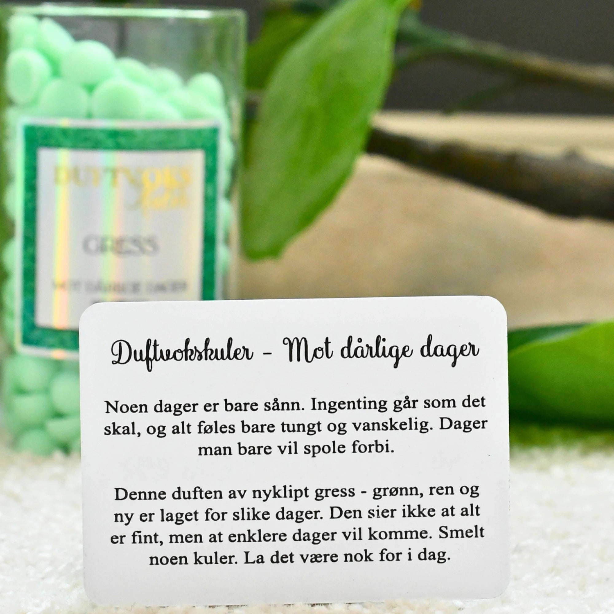 Duftvoks kulene kommer med et følelseskort mot dårlige dager. På bilde ser du kortet som følger med. I bakgrunnen står glasset med duftvoksen som fyller rommet med lukten av nyklippet gress.