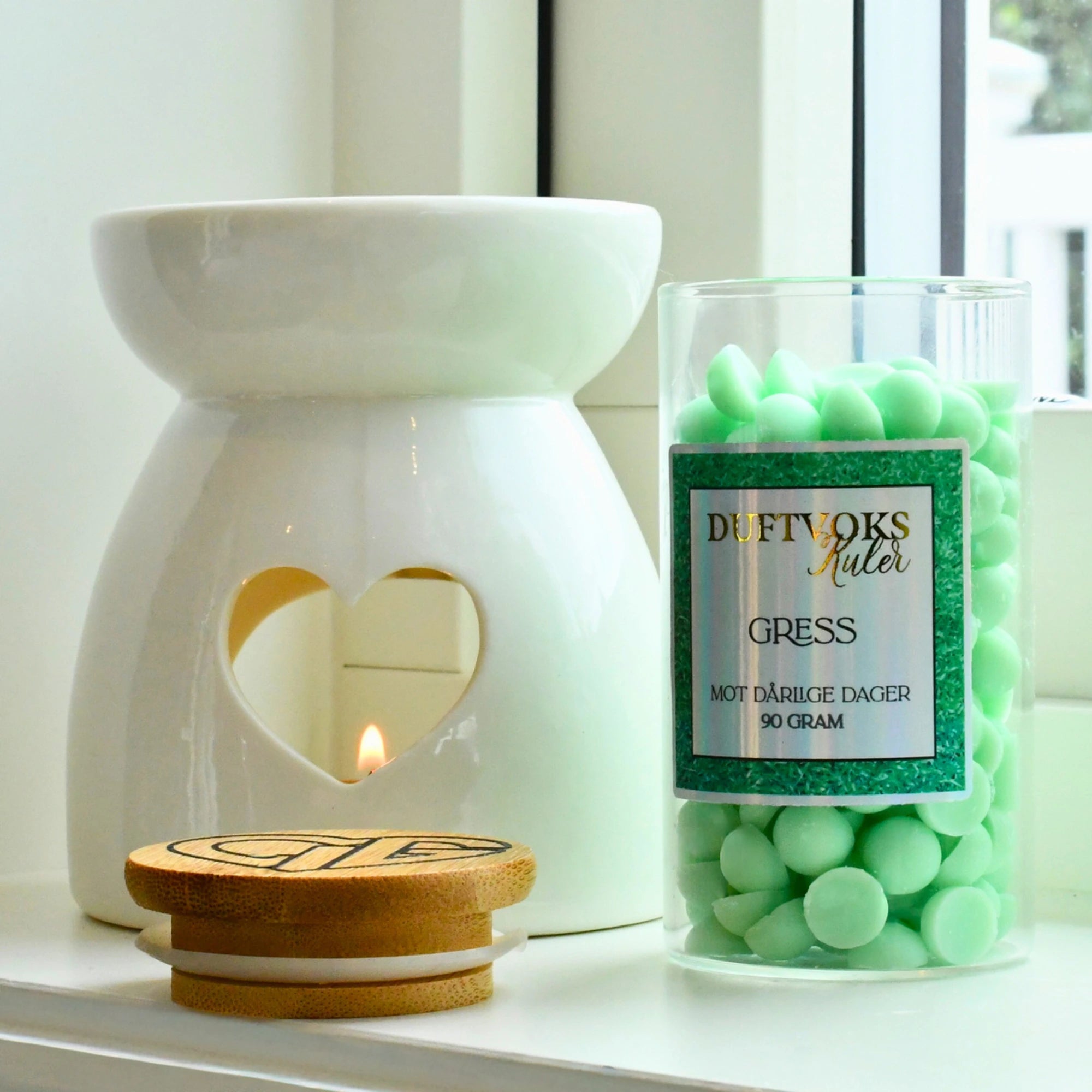 Duftvokskuler sammen med duftbrenner/voksbrenner i vindugskarmen. Duftbrenneren er tent og kulene som har en fantastisk lukt av nyklipt gress står vesidenav.