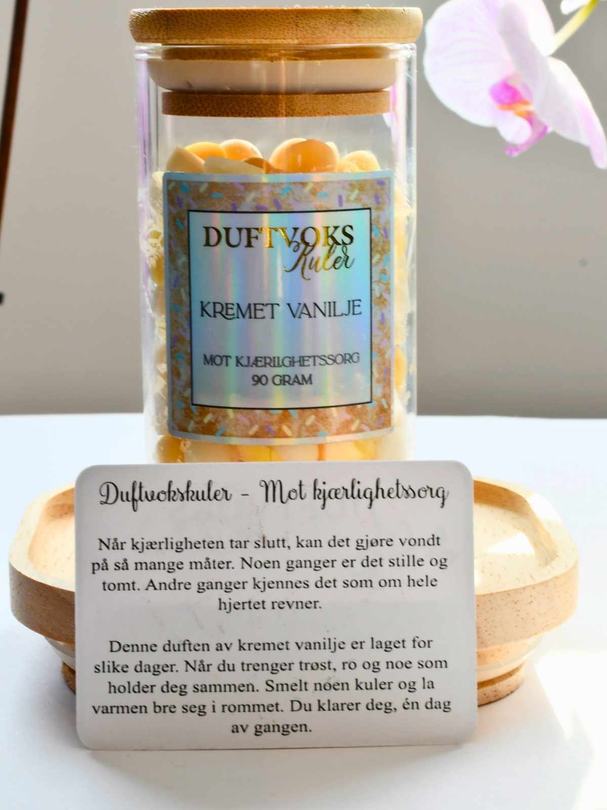 Duftvoks med duft av kremet vanlije i hvit og gul farge. Duftvoksen ligger i et glass med trelokk. En duft som skal lindre kjærlighetssorg. Foran glasset med duftvoks står et kort som beskriver duften, og hvorfor den vil virke lindrende mot kjærlighetssorg