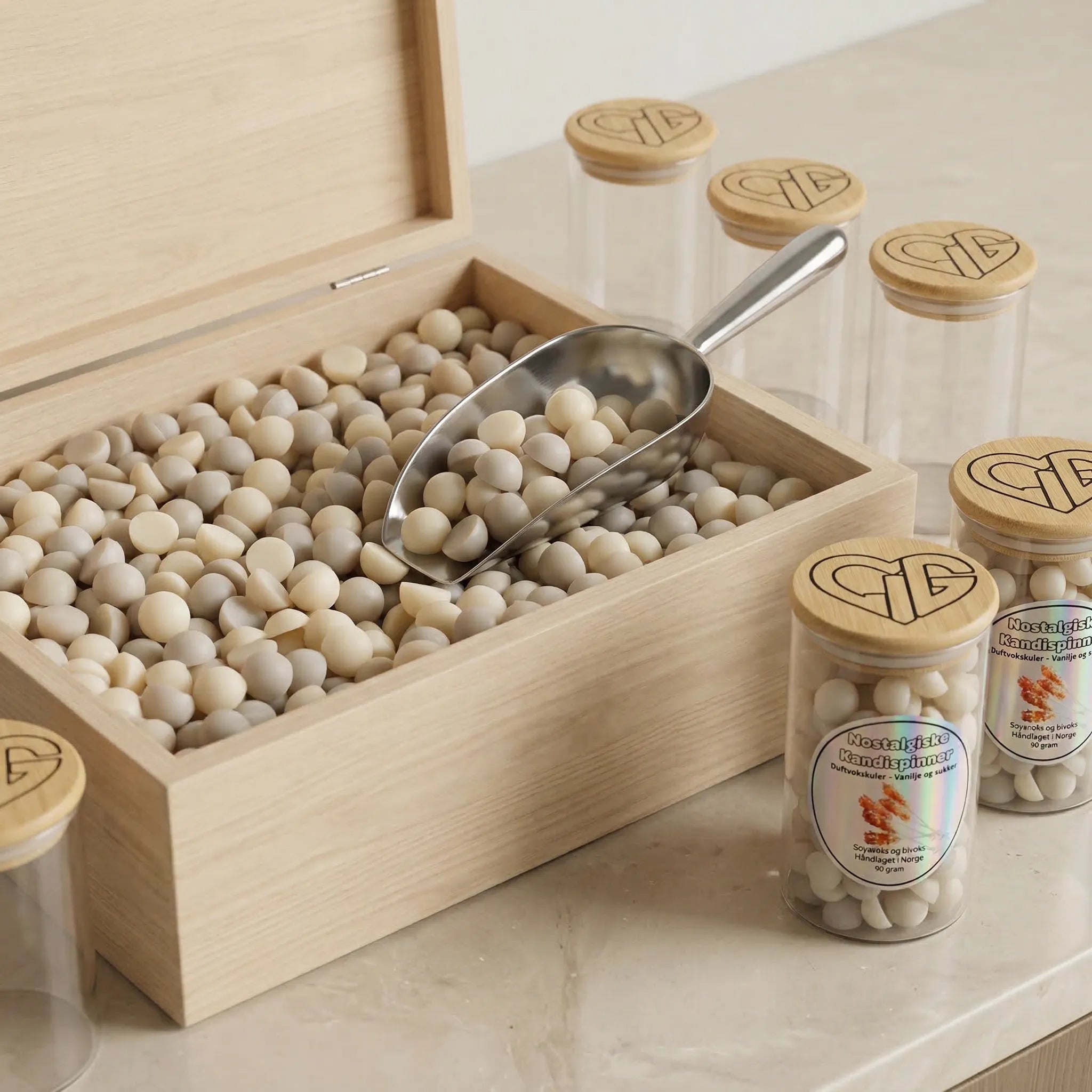 En boks fylt opp med håndlaget duftvoks. Beige og lys grå duftvokskuler med duft av nostalgiske kandispinner. Flere glass står rundt boksen, klare til å fylles opp. Glasset inneholder 90g voks, laget av 75% soyavoks og 5% bivoks. Hver kule er håndlaget i Norge.