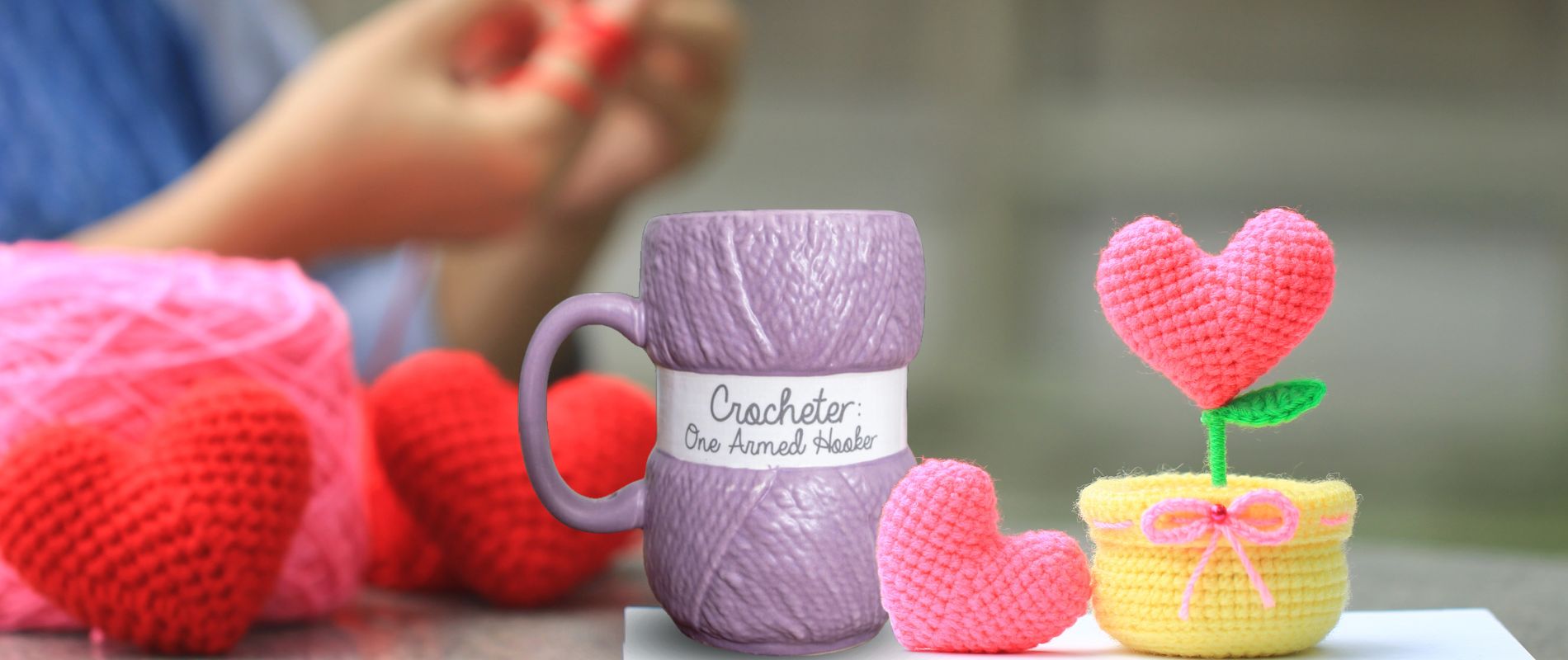 Lilla heklekopp sammen med ferdig hekla prosjekter. Masse heklede hjerter. I bakgrunnen er det en som sitter å hekler.