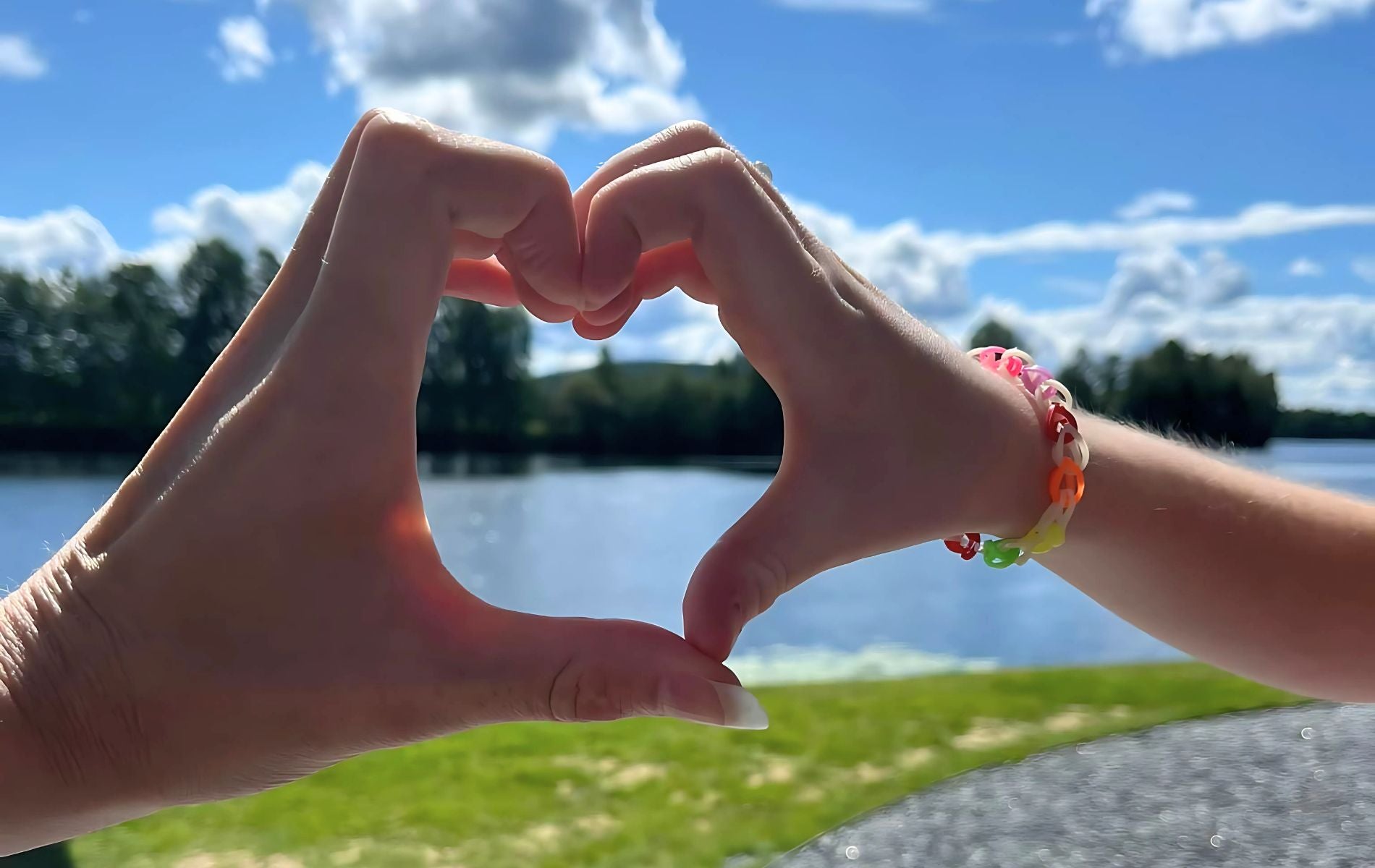 To hender som former et hjerte. En barnehånd og en voksen, som illustrerer kjærlighet. I bakgrunnen ser man et vann og en liten skog på en sommersdag