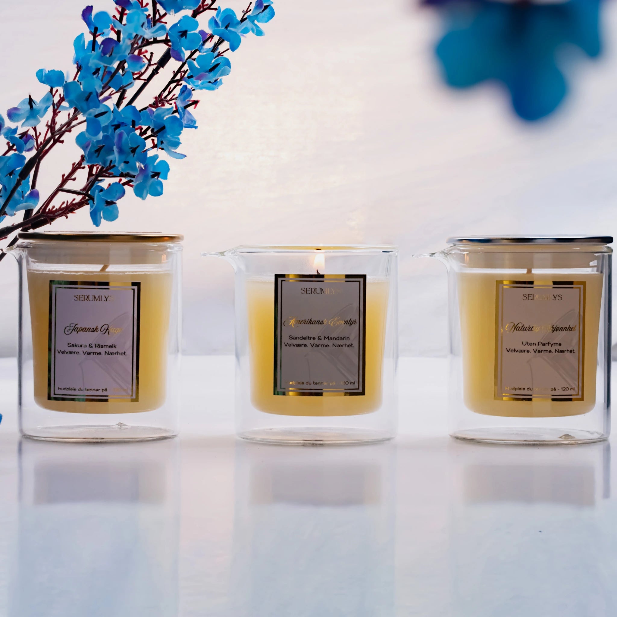 Tre glass med tre varianter av serumlys. Til venstre står den feminine Japansk hage med lokk i gull, som dufter kirsebærblomst og rismelk. I midten står den maskuline varianten med sandeltre og mandarin der lyset er tent. Til høyre står den uten parfyme med lokk i sølv. Et eksklusivt hudpleieprodukt som gir deg spa følelsen hjemme på ditt eget bad. Smelter til et næringsrikt body serum som smøres inn på kroppen mens serumet er varmt.