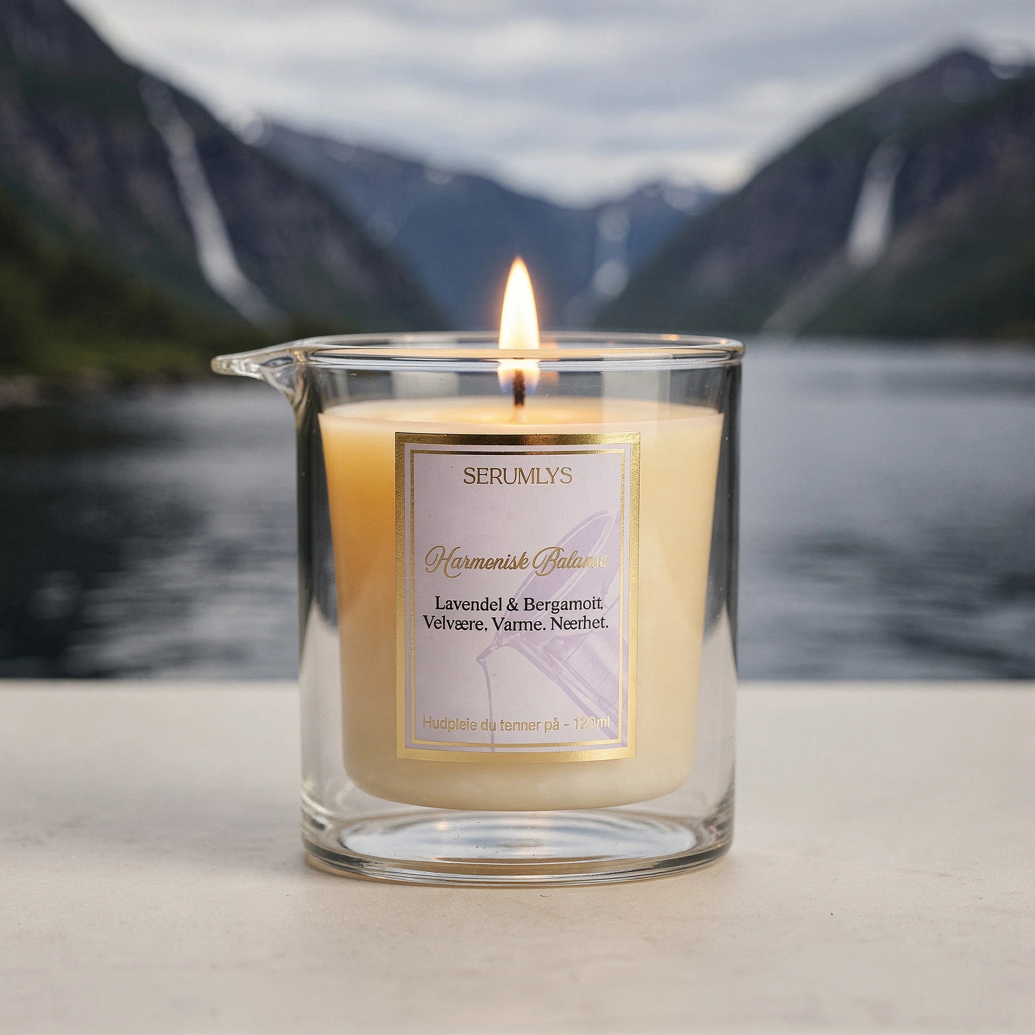 Serumlys med duft av lavendel og bergamott. Naturlig hudpleie i glass med helletut. Lyset tennes og smelter frem et pleiende kroppsserum som kan brukes på hele huden. I bakgrunnen ser man norsk natur med vann og fjell. Håndlaget kosmetikk fra Norge
