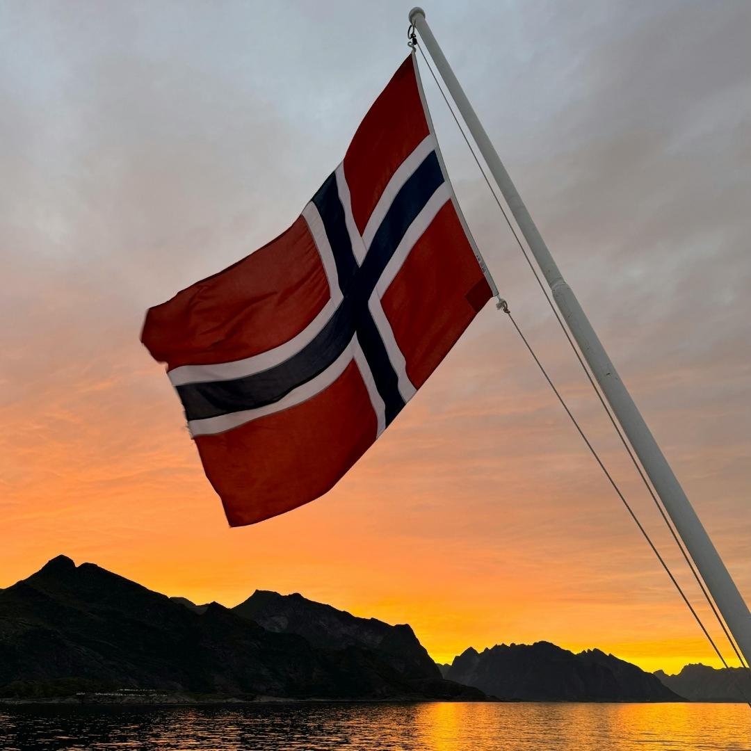Norgesflagget på stang over et vann med fjell og solnedgang. For å vise at våre produkter er utviklet og produsert av oss i Norge.