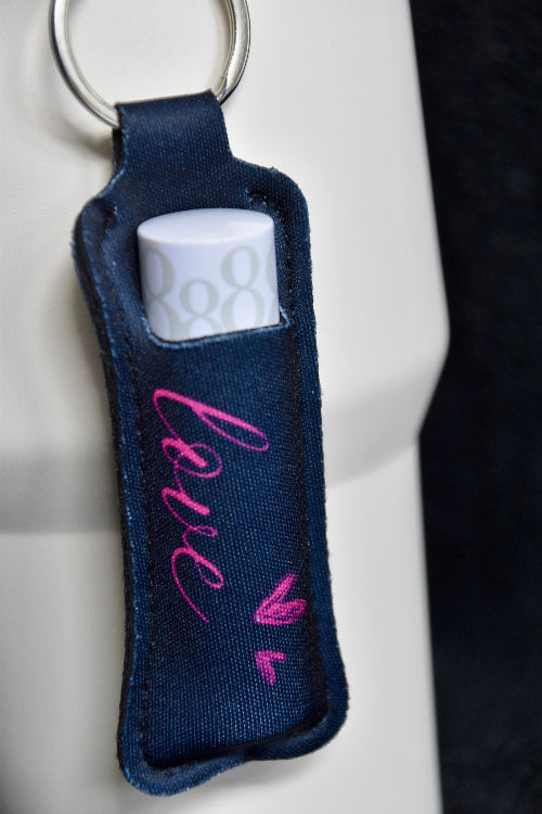 En liten veske til lipsyl eller leppestift i neopren materiale. Sort farge med rosa tekst og rosa hjerter. Designet som en nøkkelring og er festet på en beige termokopp.