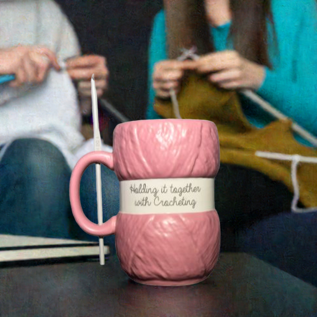 Rosa heklekopp på bordet med heklenålen i håndtaket. Mennesker i bakgrunnen som hekler