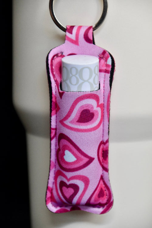 En nøkkelring med lomme til lipsyl eller leppestift. I et design med rosa hjerter. Den er festet på en beige termokopp