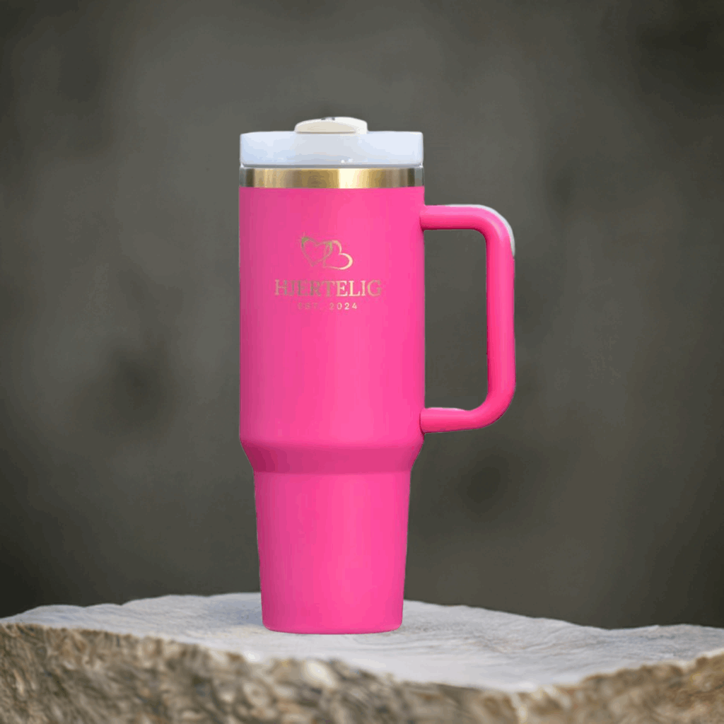 Rosa Hjertelig termokopp med gullkant og gull-logo utstilt på en stein