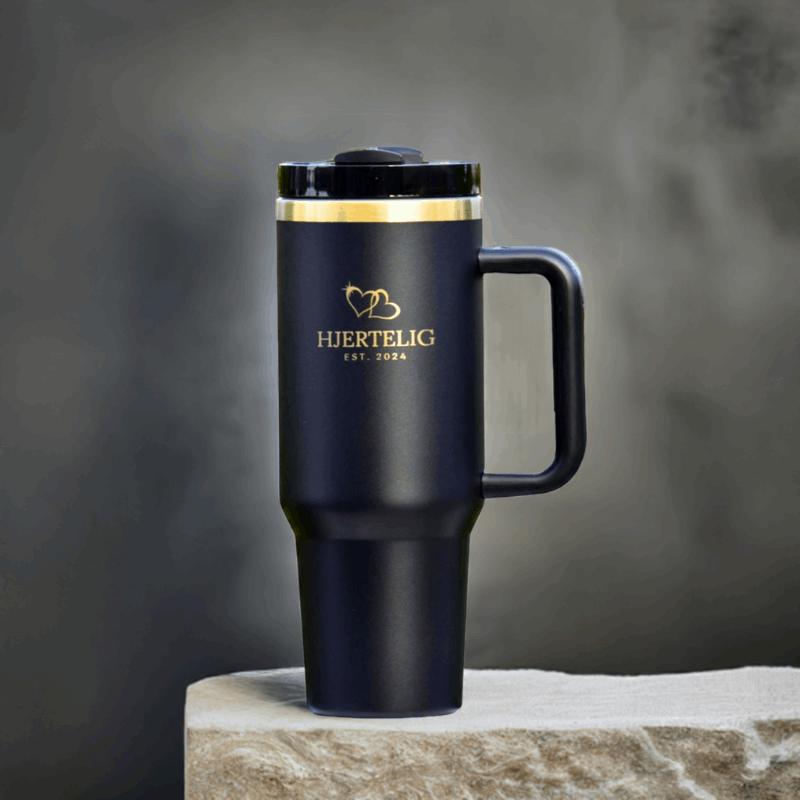 Sort termokopp med Hjertelig gull logo og gullkant utstilt på en stein. Gulldetaljene gir drikkeflasken et eksklusivt preg og er en perfekt gave til noen som sliter med å få i seg nok drikke i løpet av dagen. Lasergravert logo og gullhjerter på koppen.