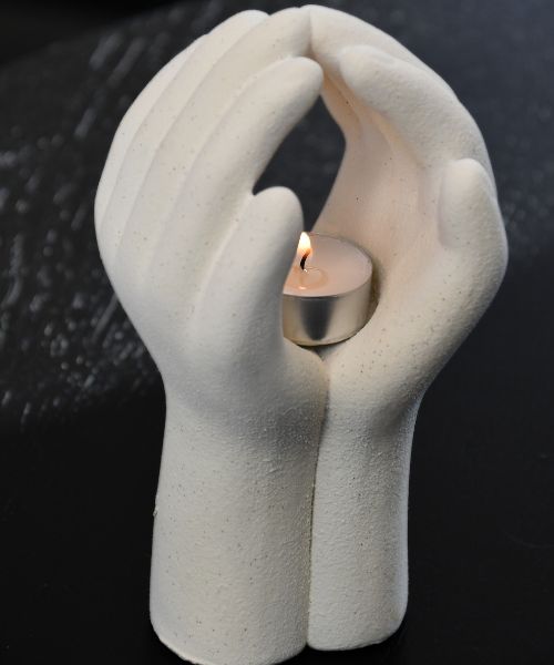 Telysholder formet som to hender som holder et telys ovenfra. Telyset er tent og man kan se flammen og lyset dette gir inni hendene. Tung keramikk telysholder som gir varme og ro.