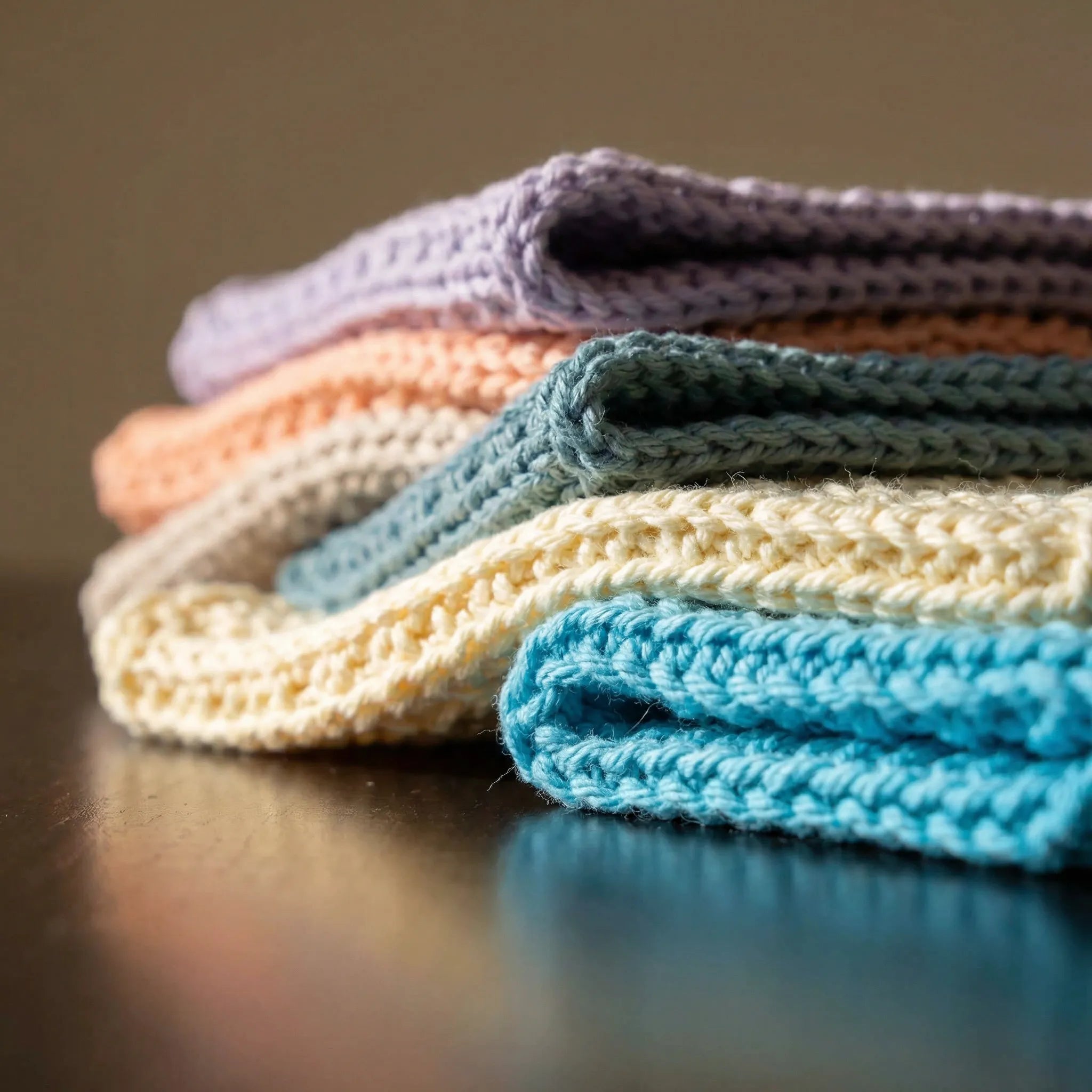 Nærbilde av vaskekluten som er strikket i 100% bomull av mamma. Her er bilde av vaske klutene i fargen turkis, lilla og gul. Detaljer av maskene og klutene som er brettet og plassert oppå hverandre. Skygger og kontraster på klutene.