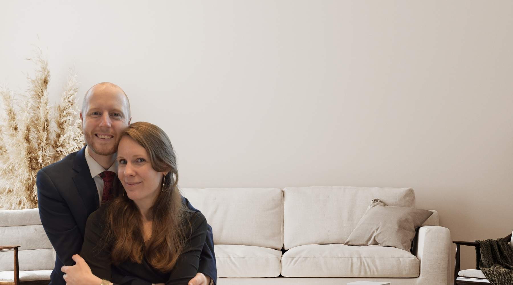 Bilde av Ståle og Siv-Helen, som er de som eier og drifter hjertelig.no. De holder rundt hverandre og smiler. I bakgrunnen står det en sofa, noen blomster og en stol