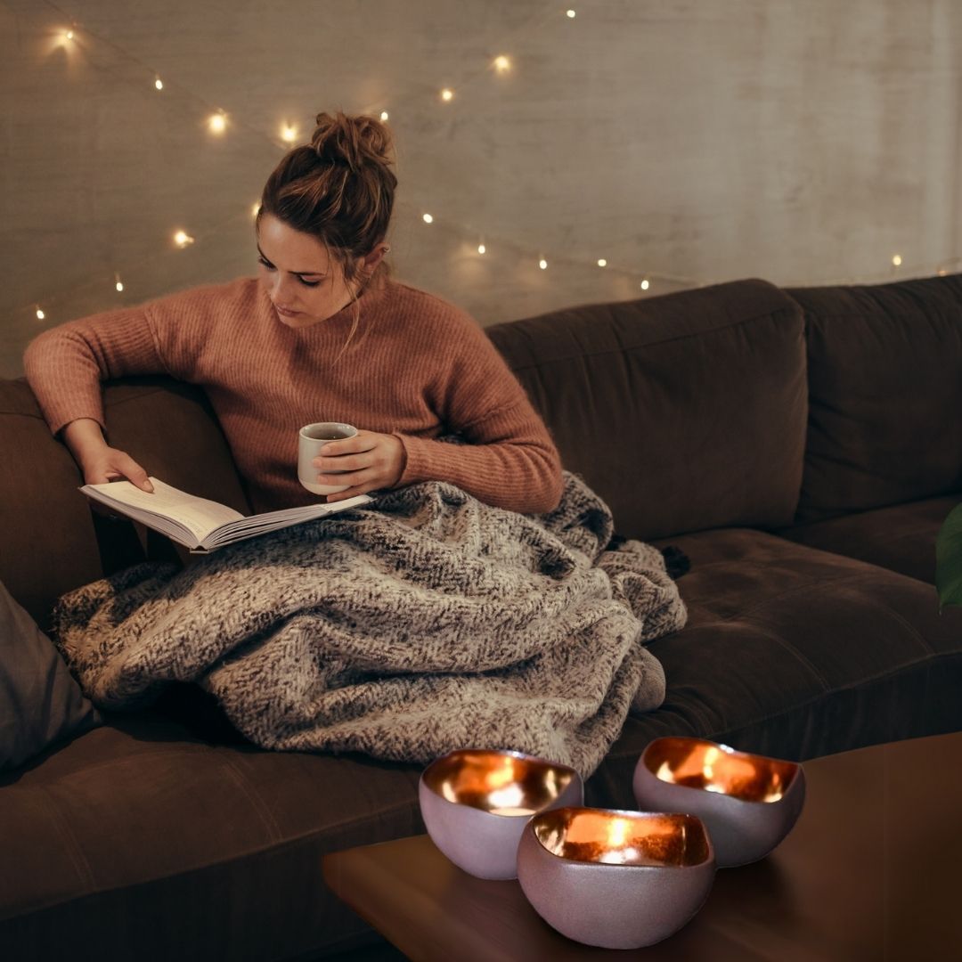 Brune telysholdere på et bord. I bakgrunnen sitter en dame i sofaen med en kopp kaffe og en bok.
