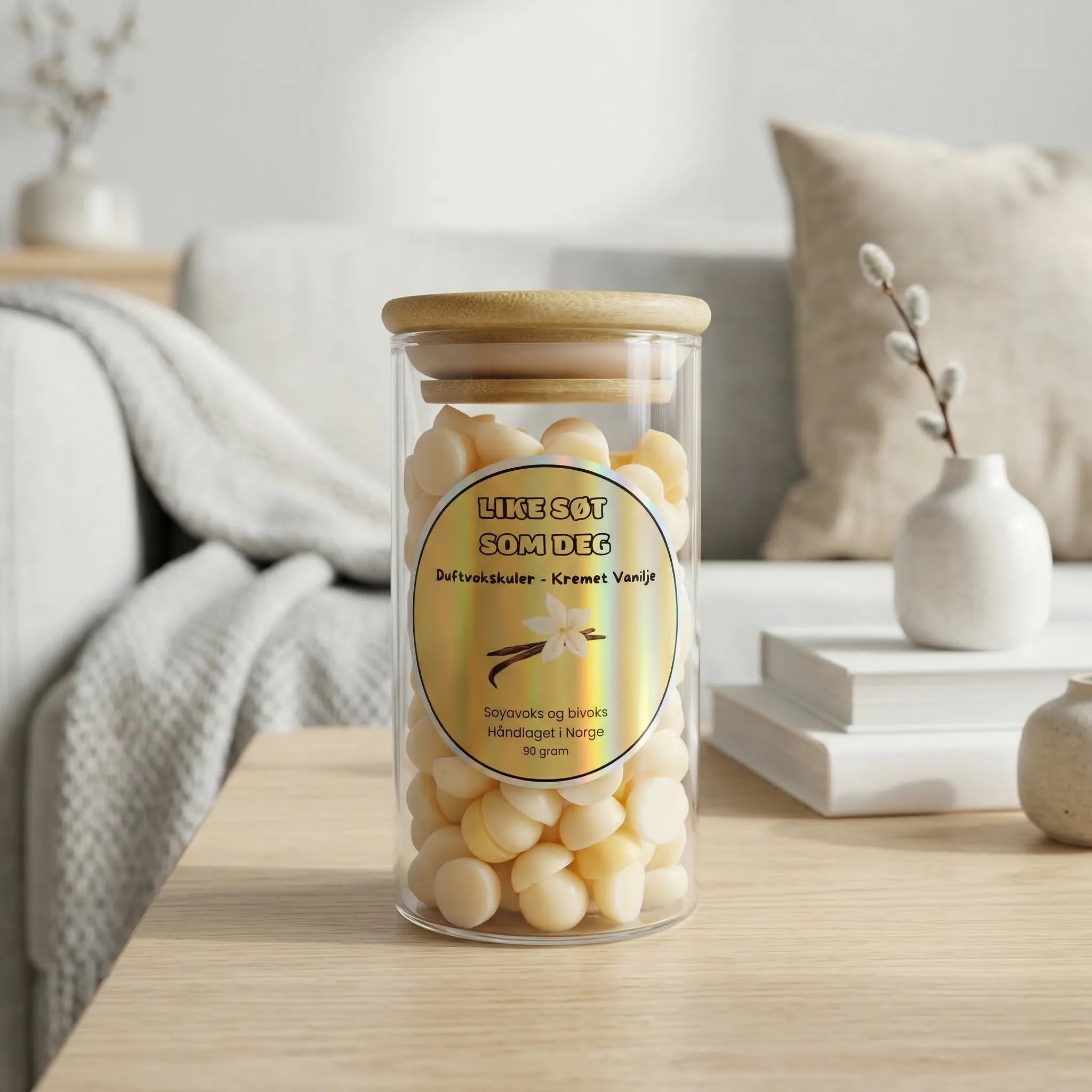 Gule duftvokskuler i glass med duft av kremet vanilje. Duften heter like søt som deg og inneholder 90g voks som er laget med soyavoks og bivoks. Glasset står på et bord i en stue. I bakgrunnen står en hvit sofa med et hvitt teppe og en beige sofapute, to bøker og to vaser.