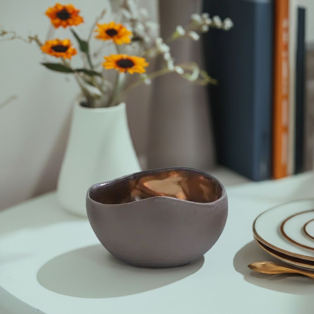 En brun håndlaget telysholder på et hvitt bord. En blomsterpotte med gule og hvite blomster i bakgrunnen