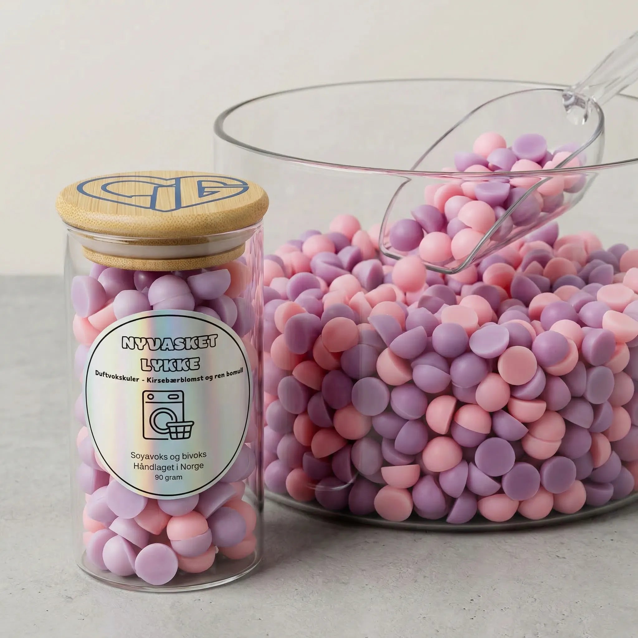 Små søte lilla og rosa duftvokskuler i en stor bolle. Glasset ved siden av er fylt opp, og rommer 90g voks. Duftvoksen er håndlaget med soyavoks og bivoks. Nyvasket lykke har en nydelig duft av nyvasket tøy.