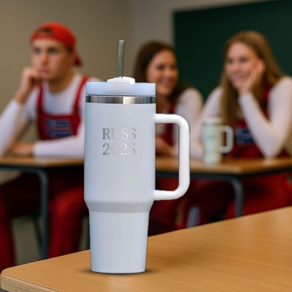 En hvit termokopp med sugerør med RUSS 2025 lasergravert. Står på en pult i et klasserom, der man ser tre russ sitte i bakgrunnen. Årets russemerch fra Hjertelig.no