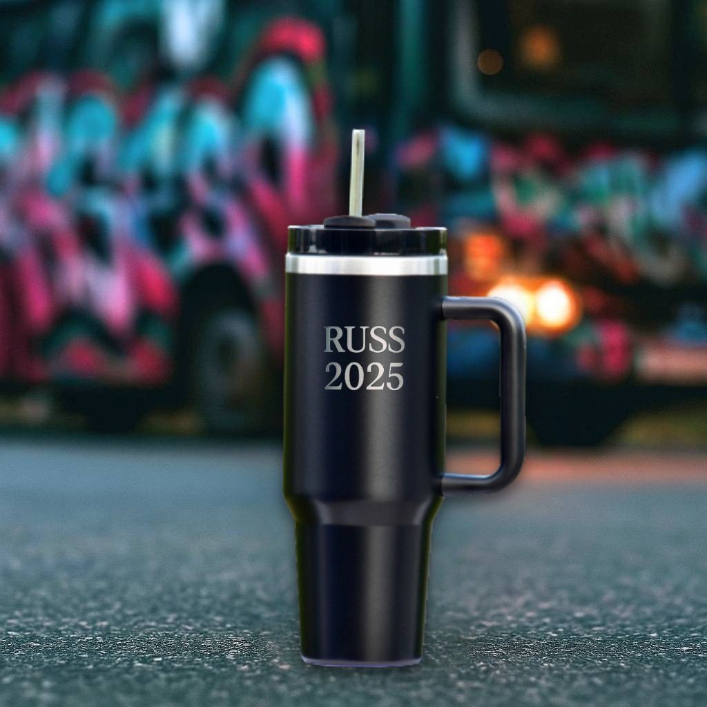 En sort termokopp med teksten russ 2025 som er lasergravert inn i termokoppen. I bakgrunnen står det en russebuss. Årets russemerch fra Hjertelig.no
