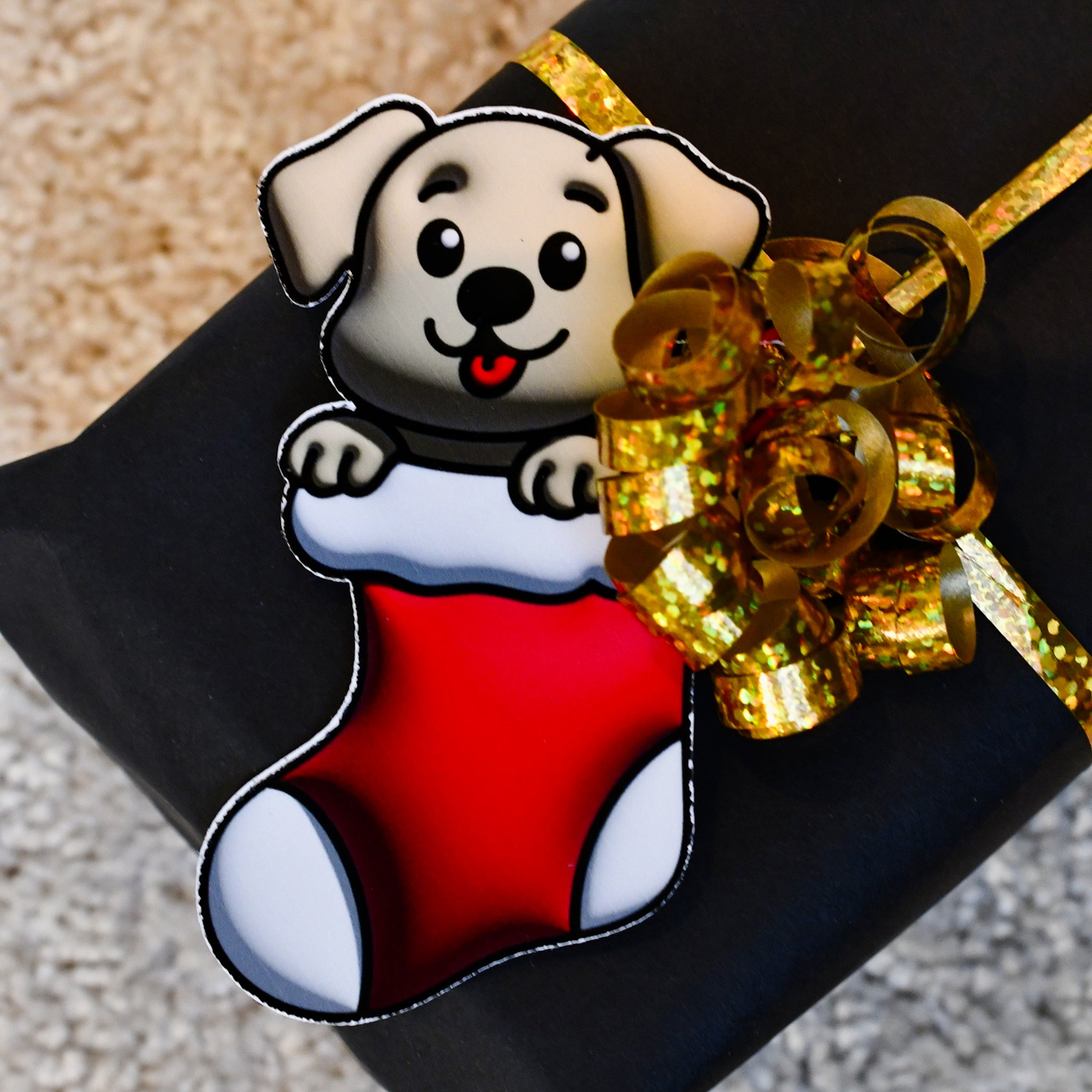 Nærbilde av den søte til fra lappen festet på julegaven med gavebåndet i gull. Gavelappen er en søt liten valp som sitter i julestrømpen og gleder seg til gavene skal bli åpnet. Gi julegavene en ekstra unik og søt detalj med disse søte lappene i år. Perfekt til både store og små.