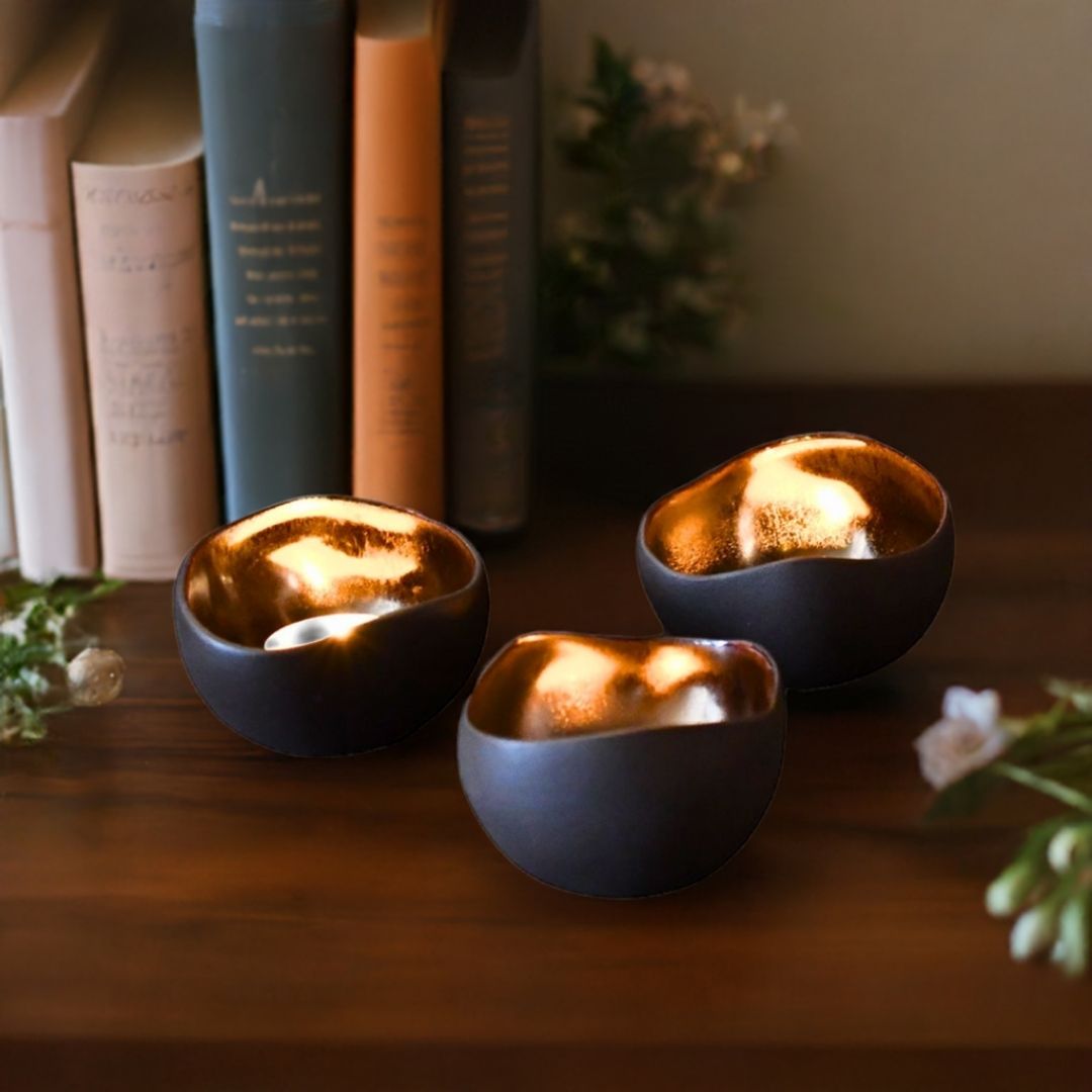 Tre håndlagde telysholdere i brun farge som står på et bord sammen med bøker og noen blomster