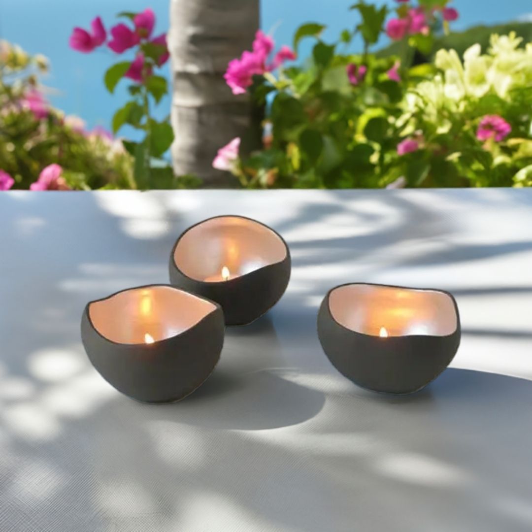 Tre telyholdere som er håndlaget. Står utendørs på et hvitt bord. I bakgrunnen ser man blomster, et tre og vann.