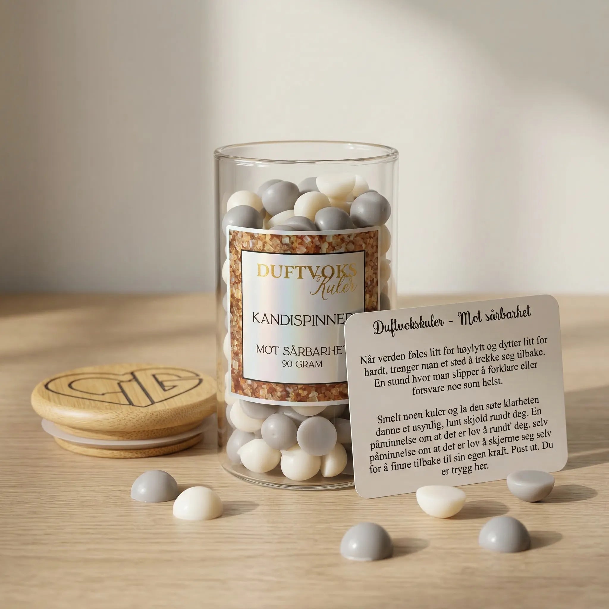Et glass med duftvoks og et kort som beskriver følelsen av sårbarhet. 90 gram voks laget av bivoks og soyavoks. Kuene er i grå og lys beige farge og dufter kandispinner. Trelokk med Hjertelig AS sin logo ligger ved siden av glasset.