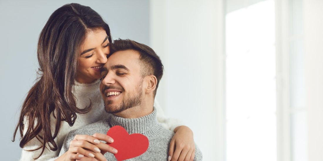 Et kjærestepar der damen står bak og holder rundt mannen. Damen holder et hjerte i papir. Begge smiler romantiskt.