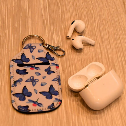 Veske til airpods som ligger på bakken med sommerfuglmotiv vesidenav ligger airpods som passer i vesken. 
