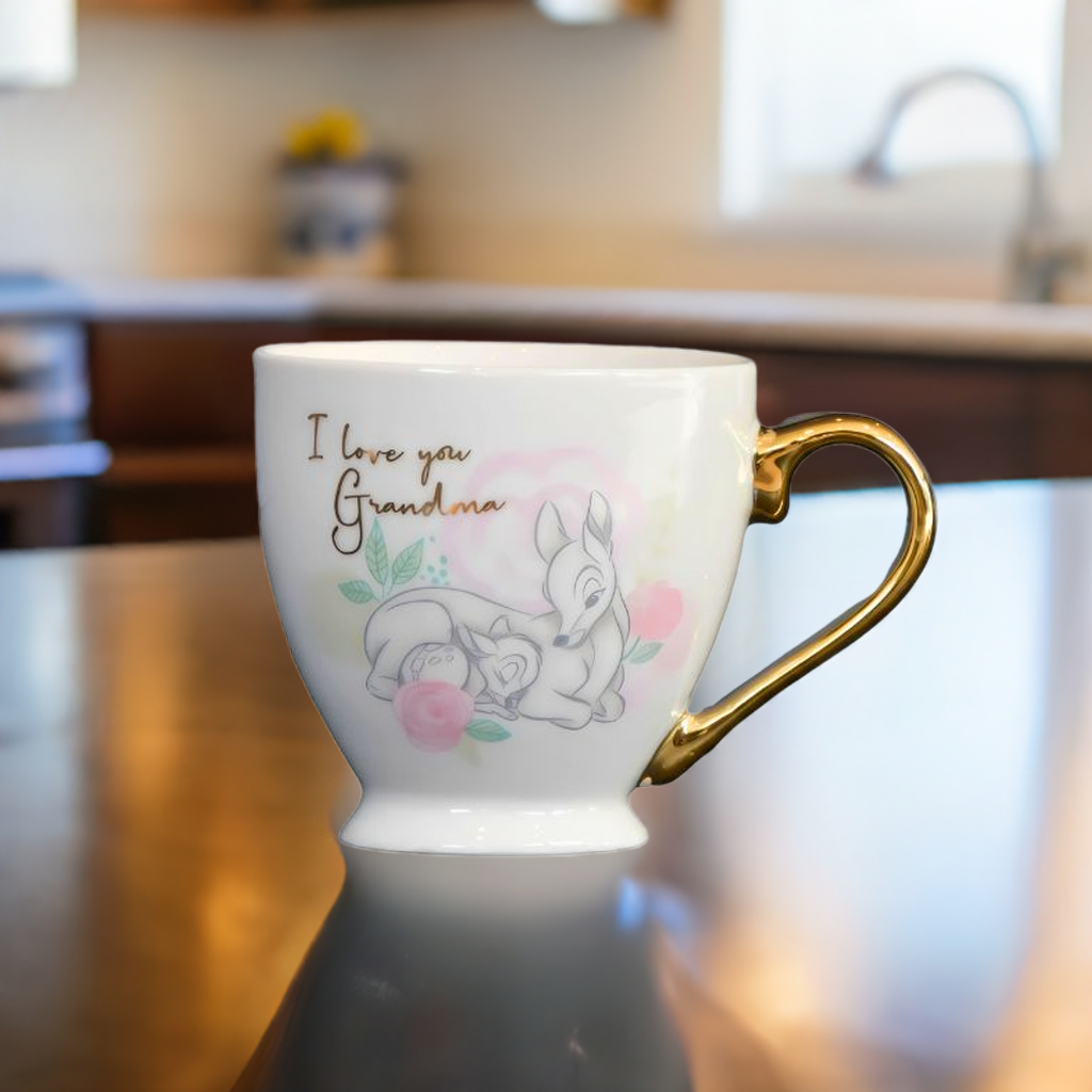En Disney kopp med bambi motiv og gullhåndtak som er formet som et halvt gullhjerte. Kruset står på et kjøkkenbord. I bakgrunnen kan man se kjøkkenkran og gule blomster. En perfekt julegave eller bursdagsgave til din bestemor. Koppen har et nydelig design av bambi med teksten I love you grandma.