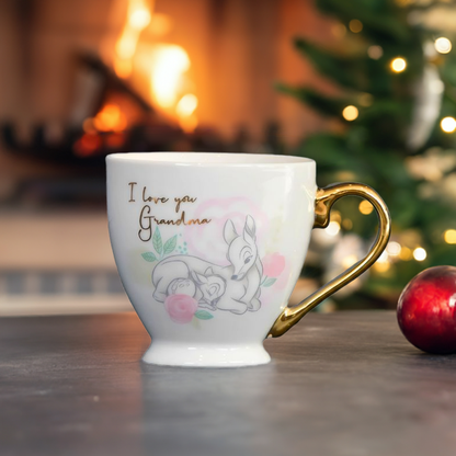 Kopp med bambi motiv til grandma. Perfekt gave til jul. Her er juletreet i bakgrunnen med en rød julekule til høyre for kruset. Fint design av bambi fra Disney på en hvit kopp med et gullhåndtak formet som et halvt hjerte. En perfekt gave til din bestemor for å vise hvor mye hun betyr for deg.