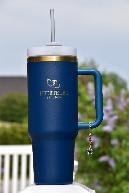 Et smykke med sommerfugl motiv som er festet rundt håndtaket på en blå tumbler med gullkant og logo