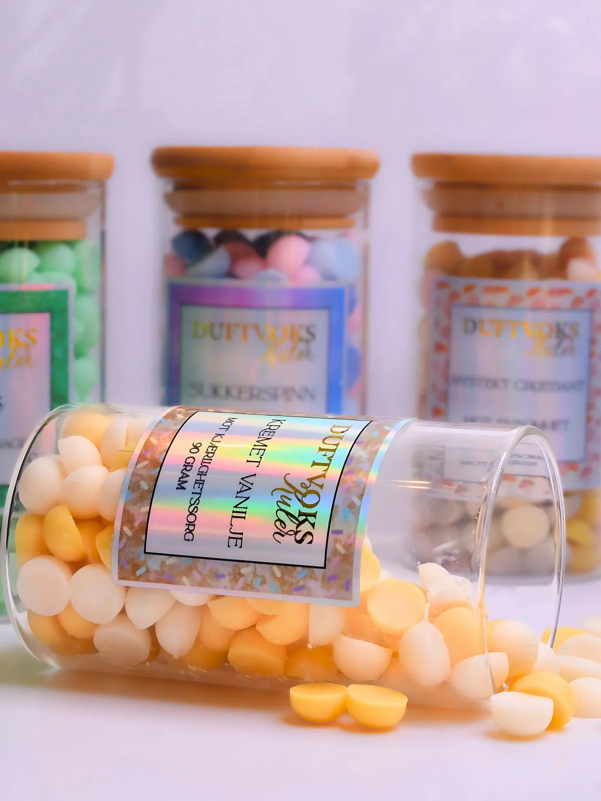 Kremet vanilje duftvoks laget av soya voks. Her ligger glasskrukken med noen duftvoks kuler rundt seg. I bakgrunnen kan du se tre av våre andre fantastiske dufter i vår duftvoks serie for følelser. 