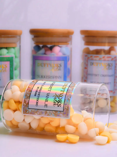 Kremet vanilje duftvoks laget av soya voks. Her ligger glasskrukken med noen duftvoks kuler rundt seg. I bakgrunnen kan du se tre av våre andre fantastiske dufter i vår duftvoks serie for følelser. 