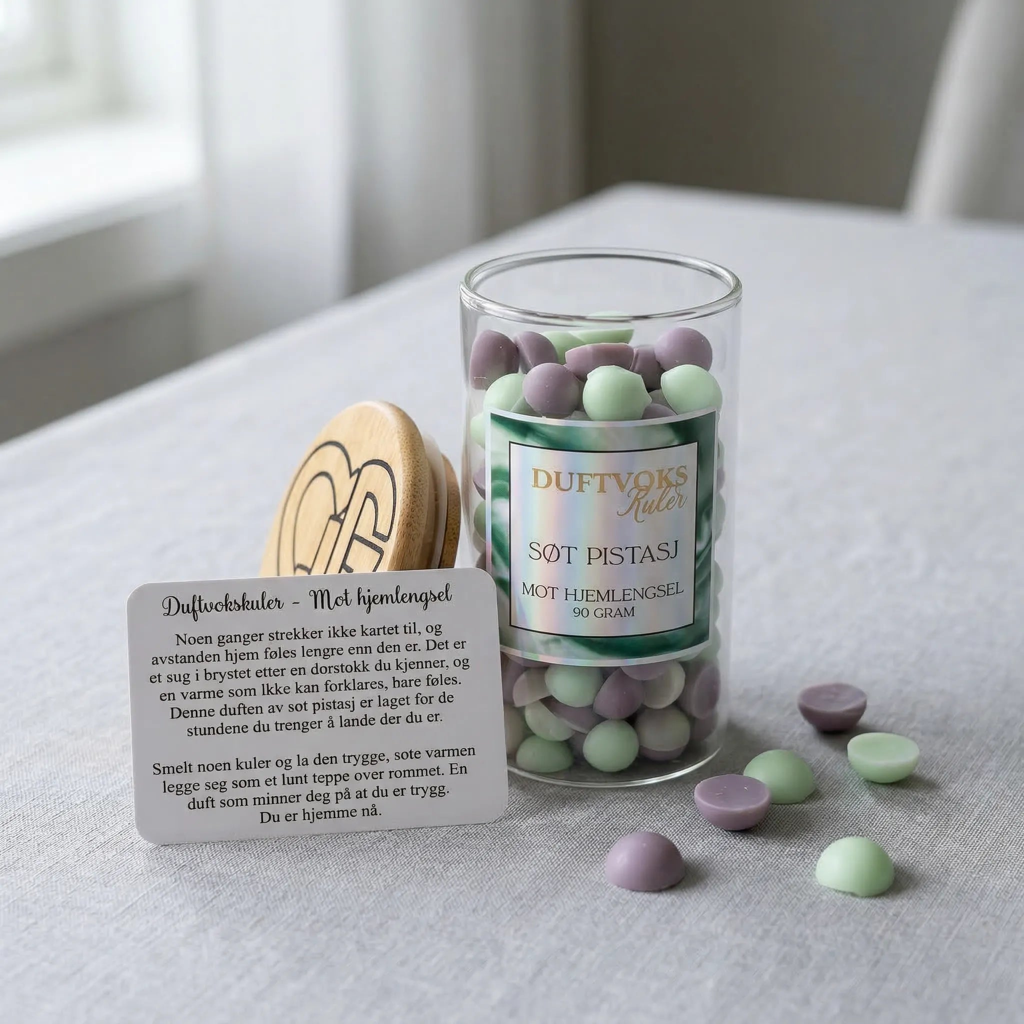 Et glass med duftvoks som lukter søt pistasj. Ved siden av glasset står et kort som beskriver hvorfor denne duften kan virke lindrende mot hjemlengsel. En av flere dufter i serien "sett en duft på dine følelser" håndlaget i norge med bivoks og soyavoks.