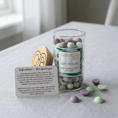Et glass med duftvoks som lukter søt pistasj. Ved siden av glasset står et kort som beskriver hvorfor denne duften kan virke lindrende mot hjemlengsel. En av flere dufter i serien "sett en duft på dine følelser" håndlaget i norge med bivoks og soyavoks.
