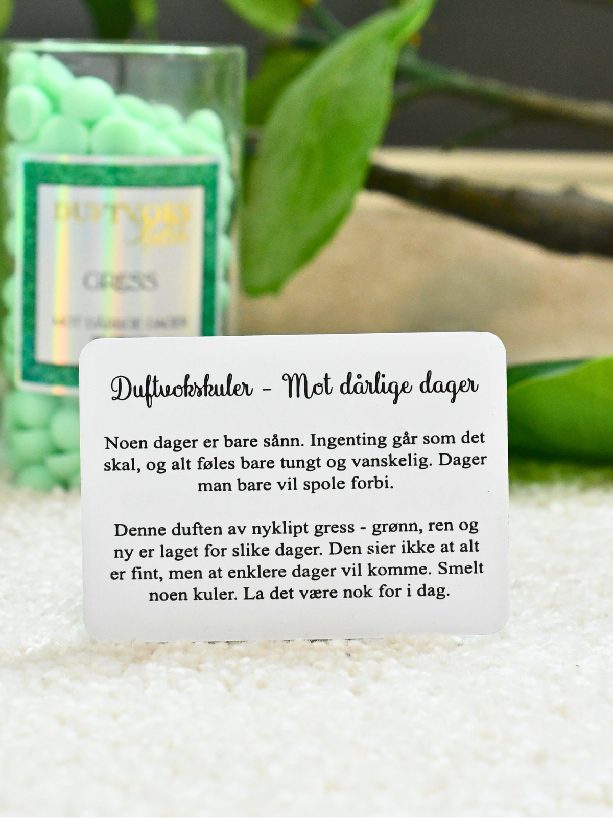 Duftvoks kulene kommer med et følelseskort mot dårlige dager. På bilde ser du kortet som følger med. I bakgrunnen står glasset med duftvoksen som fyller rommet med lukten av nyklippet gress. 