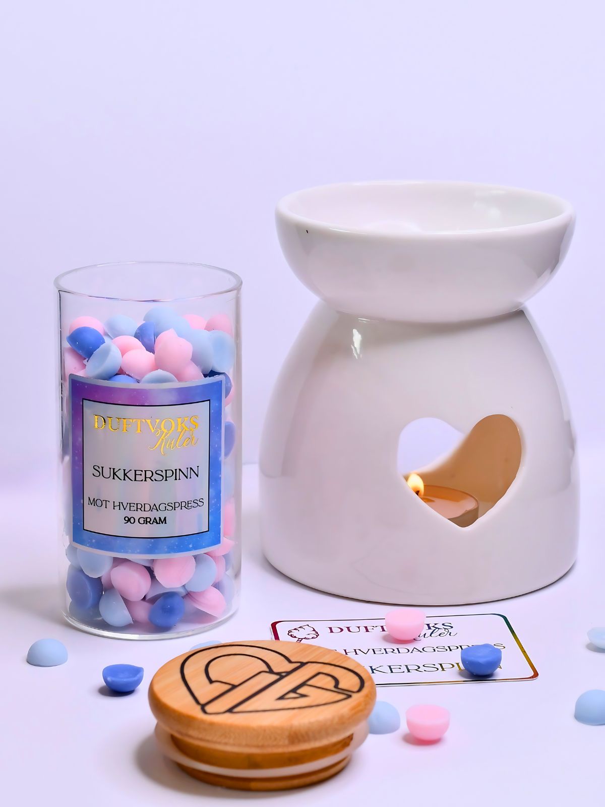 Duftvoks kuler i bruk i duftbrenner med hverte. Duftvoks kulene ligger rundt, og bambuslokket er tatt av glasskrukken. Glasset inneholder 90gram med soyavoks kuler i rosa og blå farge. Man kan se hvertelig logoen om er på lokket. 