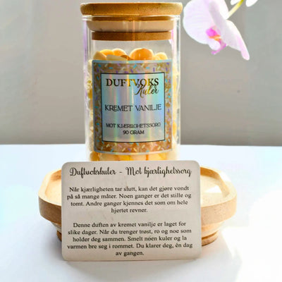 Duftvoks med duft av kremet vanlije i hvit og gul farge. Duftvoksen ligger i et glass med trelokk. En duft som skal lindre kjærlighetssorg. Foran glasset med duftvoks står et kort som beskriver duften, og hvorfor den vil virke lindrende mot kjærlighetssorg