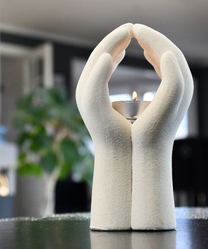 Telys i midten av to hender. Telysholderen har telyset i midten av hendene og står på et bord. Her ser man hvordan denne vil se ut i stuen. Flammen på telyset som lyser opp inni hendene. Unikt design til ditt hjem, som gir en følelse av ro og fred. Fargen på keramikk holderen er beige. 