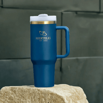 Nydelig blå termokopp med gull logo og gullkant som står utstilt på en stein