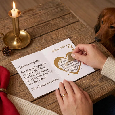 Kreativ julegave i et dachshund julekort. Overraskelsen ligger under skrapehjertet. Spenning og underholdning når man skal finne ut hva som befinner seg under hjertet ved å skrape bort klistremerke med en mynt. Her ser man julekortet i brukt. Dachshunden ser spent på overraskelsen mens mamma skraper den fram. 