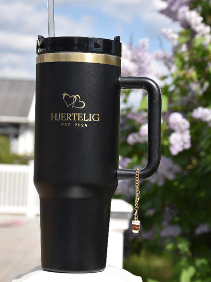 Et kaffekopp smykke som henger på en sort termokopp med gullkant og Hjertelig logo