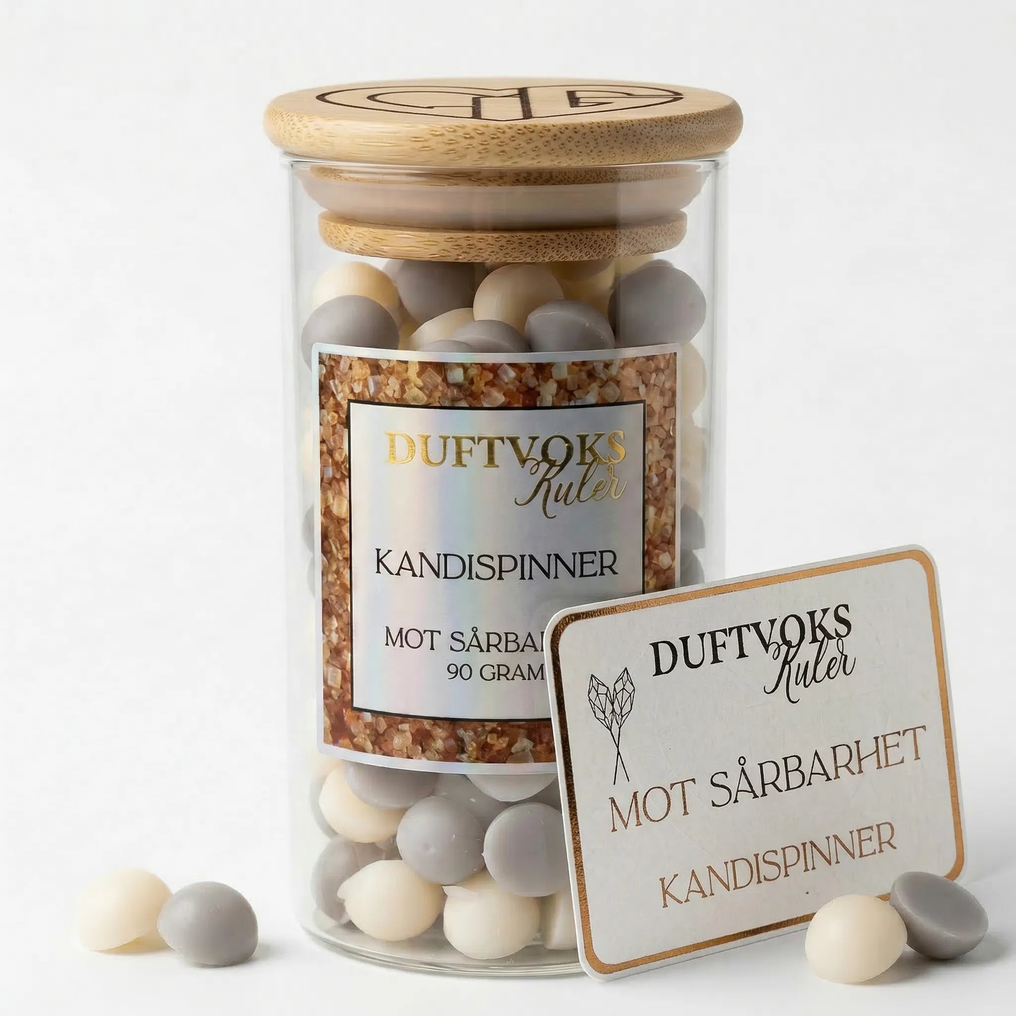 Håndlaget duftvoks fra Norge med duft av kandispinner. En duft som skal virke lindrende mot følelsen av sårbarhet. 90g med voks laget av bivoks og soyavoks i t glass med trelokk. Ved siden av glasset står følelseskortet som følger med produktet, som forklarer hvorfor denne duften kan virke lindrende mot det å være ekstra sårbar. Duftvokskulene er grå og lys beige. 