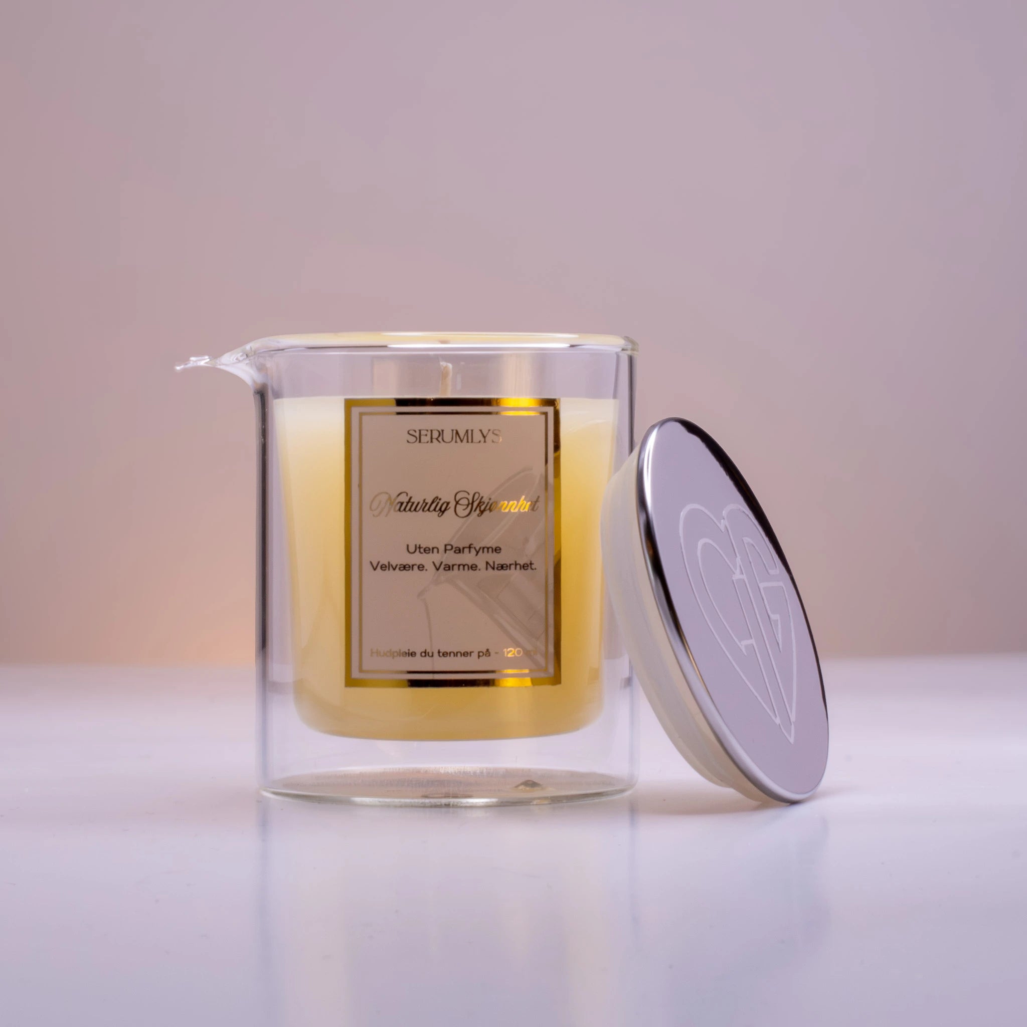 Et serumlys i glass med helletut. Ved siden av glasset står et lokk i sølv med Hjertelig AS sin logo utenpå. Lyset er et hudpleieprodukt med naturlige oljer. Når lyset smelter så helles oljene på kroppen rett fra glasset.