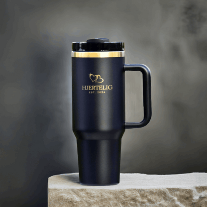 Sort termokopp med Hjertelig gull logo og gullkant utstilt på en stein. Gulldetaljene gir drikkeflasken et eksklusivt preg og er en perfekt gave til noen som sliter med å få i seg nok drikke i løpet av dagen. Lasergravert logo og gullhjerter på koppen. 
