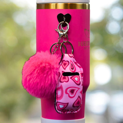 Svart krok på rosa termokopp. På kroken er det festet rosa hjerte veske til airpods og en rosa dusk. 
